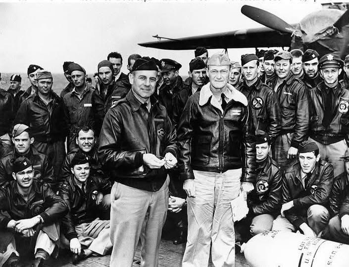 Jimmy Doolittle and Captain Marc Mitscher stand on the flight deck of the USS Hornet surrounded by Raiders in leather flight jackets