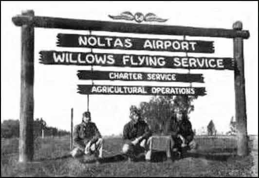 Three men in flight gear sit under a large wooden sign reading Noltas Airport, Willows Flying Service, Charter Service, Agricultural Operations
